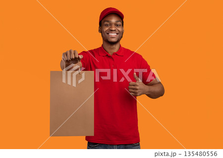 Portrait of african american delivery man in red uniform holding craft paper bag, showing thumbs up gesture, isolated on orange background. Design, mockup, blank space on package for advert 135480556