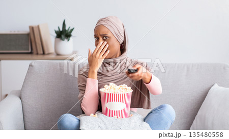 Young black woman in hijab watching scary movie, covering her mouth in horror, eating popcorn on sofa at home. Millennial Muslim lady feeling afraid, switching thriller off with remote control Young black woman in hijab watching scary movie, covering her mouth in horror, eating popcorn on sofa at home. Millennial Muslim lady feeling afraid, switching thriller off with remote control 135480558