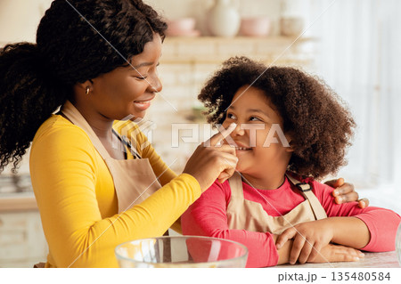 Loving Black Mom Playing With Her Daughter While They Baking In Kitchen, Cheerful African American Woman And Little Curly Girl Wearing Aprons And Having Fun Together, Enjoying Making Homemade Pastry Loving Black Mom Playing With Her Daughter While They Baking In Kitchen, Cheerful African American Woman And Little Curly Girl Wearing Aprons And Having Fun Together, Enjoying Making Homemade Pastry 135480584