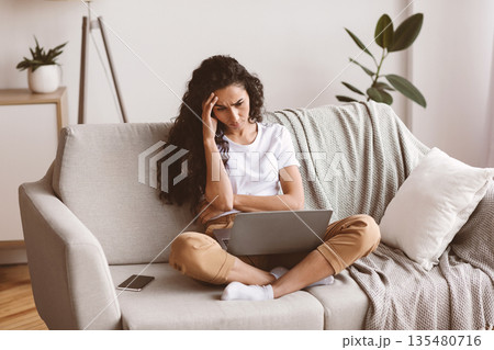 Upset young jobless woman sitting on couch with laptop on her lap, touching her head and looking at screen, home interior. Thoughtful curly dark-haired lady having financial difficulties, copy space Upset young jobless woman sitting on couch with laptop on her lap, touching her head and looking at screen, home interior. Thoughtful curly dark-haired lady having financial difficulties, copy space 135480716