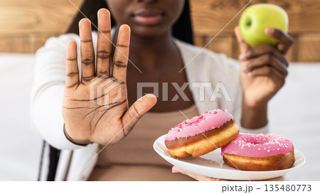 Healthy Eating During Pregnancy. Black Pregnant Lady Refusing Unhealthy Donuts And Choosing Apple For Snack, Showing Stop Gesture With Palm, Sitting On Bed At Home, Closeup Shot With Selective Focus Healthy Eating During Pregnancy. Black Pregnant Lady Refusing Unhealthy Donuts And Choosing Apple For Snack, Showing Stop Gesture With Palm, Sitting On Bed At Home, Closeup Shot With Selective Focus 135480773