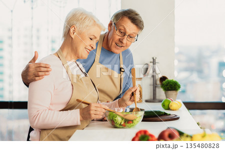 Side View Of Happy Senior Couple Cooking In Kitchen, Making Fresh Vegetable Salad For Dinner Together Standing Indoors. Aged Spouses Preparing Food At Home. Older Family Weekend Leisure 135480828