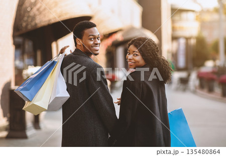 Sale, discounts and Black Friday at city mall before holidays. Smiling young african american guy and woman in coats with colored bags turns and looks at camera on street in november, copy space Sale, discounts and Black Friday at city mall before holidays. Smiling young african american guy and woman in coats with colored bags turns and looks at camera on street in november, copy space 135480864