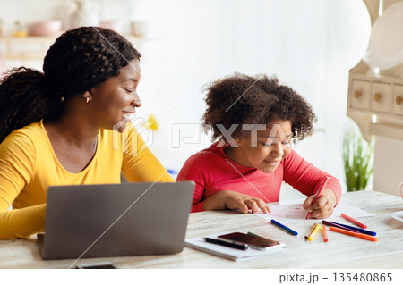 Cute Little Black Girl Drawing In Kitchen Next To Her Mom Working Online On Laptop. Young African American Mother Sitting At Table With Computer Spending Time With Her Kid And Enjoying Freelance Work Cute Little Black Girl Drawing In Kitchen Next To Her Mom Working Online On Laptop. Young African American Mother Sitting At Table With Computer Spending Time With Her Kid And Enjoying Freelance Work 135480865