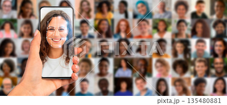 A person holds a smartphone displaying a facial recognition scan in a busy environment. Behind, a collage of diverse faces illustrates modern identification technology's extensive application. A person holds a smartphone displaying a facial recognition scan in a busy environment. Behind, a collage of diverse faces illustrates modern identification technology's extensive application. 135480881
