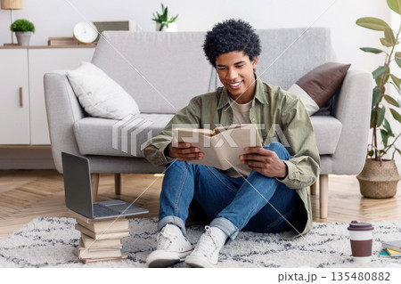 Full length portrait of young black guy with book and laptop having webinar, attending online class from home. Teenage student studying remotely during covid-19 pandemic 135480882
