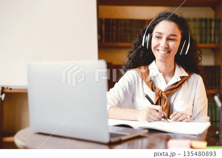 Distance Education. Smiling woman in wireless headset sitting at desk, using laptop and writing in notebook. Lady watching tutorial, lecture or webinar, studying online at home. E-learning 135480893