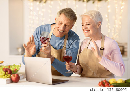 Joyful Senior Couple At Laptop Celebrating Christmas Making Video Call Toasting Holding Wine Glasses In Kitchen At Home Decorated And Illuminated With Lights. Modern Christmas And New Year Celebration 135480905