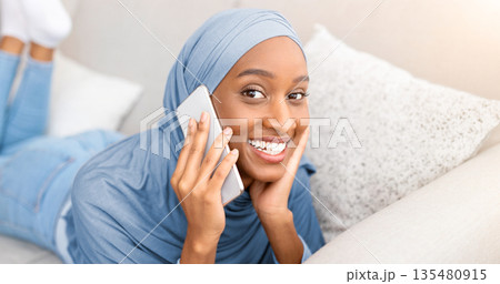 Attractive young Muslim woman in hijab chatting on cellphone, having pleasant talk with friend while lying on sofa at home. Modern technologies and communication concept 135480915