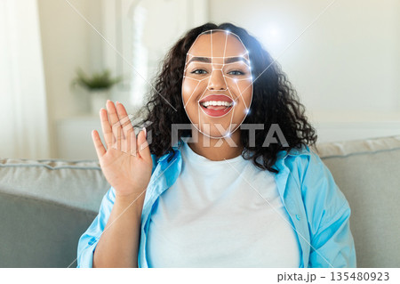 A person with curly hair smiles while waving, engaging in a facial recognition identification process. The modern setting reflects the integration of technology into everyday life. 135480923