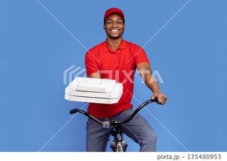 Best Fast Food Delivery. Portrait of smiling african courier wearing red cap and uniform holding pizza boxes from restaurant and riding bicycle, isolated on blue studio background 135480953