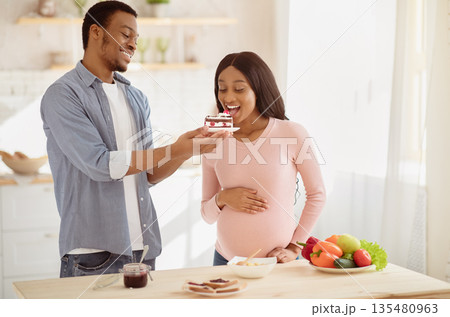 Positive African American guy offering tasty cake to his pregnant girlfriend at home. Attractive black man tempting his expectant wife with yummy dessert in kitchen. Poor pregnancy diet concept Positive African American guy offering tasty cake to his pregnant girlfriend at home. Attractive black man tempting his expectant wife with yummy dessert in kitchen. Poor pregnancy diet concept 135480963