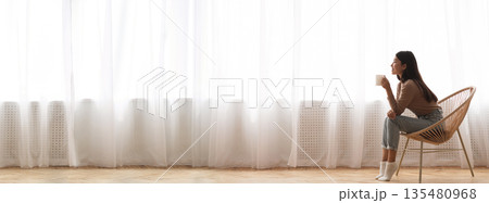 Millennial Asian Girl Relaxing In Modern Chair, Sitting With Cup Of Coffee In Front Of Window, Side View Panorama With Empty Space, Long Wide Banner 135480968