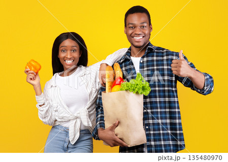 Healthy eats, modern ordering products online. Happy young african american husband and wife hold paper bags with vegetables and showing thumb up and orange pepper, isolated on yellow background Healthy eats, modern ordering products online. Happy young african american husband and wife hold paper bags with vegetables and showing thumb up and orange pepper, isolated on yellow background 135480970