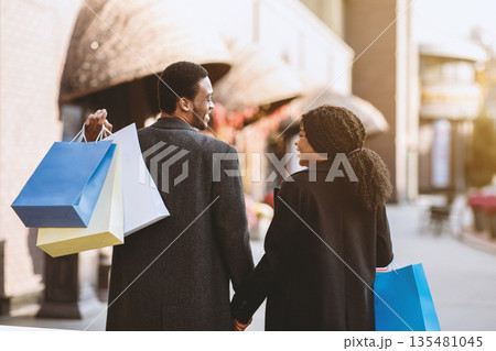 Autumn time and black friday, season sale and holiday. Happy young african american man and woman with lots of colorful paper bags walking to street after shopping and look at each other, back 135481045