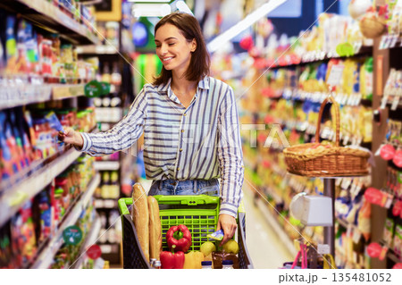 Everyday Shopping Concept. Smiling Young Housewife Pushing Trolley Cart Between Aisles In Grocery Store. Positive Woman Buying Food, Taking Products From Shelf. Consumerism And Lifestyle Everyday Shopping Concept. Smiling Young Housewife Pushing Trolley Cart Between Aisles In Grocery Store. Positive Woman Buying Food, Taking Products From Shelf. Consumerism And Lifestyle 135481052
