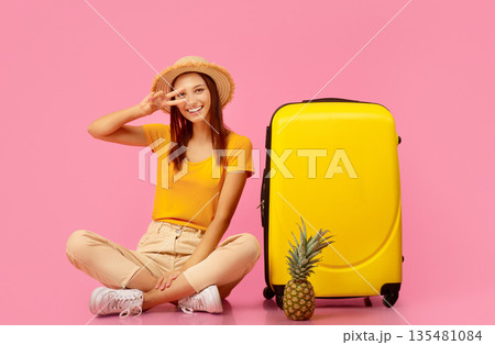 Happy woman in summer clothes with pineapple showing peace gesture, sitting by suitcase, posing on pink studio background. Carefree young lady travelling to exotic warm countries, hot deals 135481084