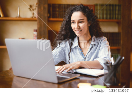 Doing Project. Portrait of curly young woman using laptop computer and typing on keyboard, writer sitting at table. Female manager working remotely or studying online at home, telecommuting Doing Project. Portrait of curly young woman using laptop computer and typing on keyboard, writer sitting at table. Female manager working remotely or studying online at home, telecommuting 135481097