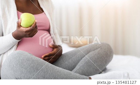 Black pregnant woman eating green apple while sitting on bed at home, enjoying healthy nutrition during pregnancy, having vitamin snack while resting in bedroom, cropped image with free space 135481127