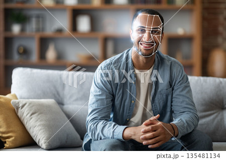 African American man sits comfortably on a sofa in a cozy living space while facial recognition technology scans his face for identification purposes. African American man sits comfortably on a sofa in a cozy living space while facial recognition technology scans his face for identification purposes. 135481134