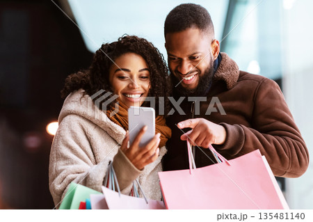 Sales, Offers And Discount. Smiling african american family of two using mobile phone, guy pointing at gadget, couple standing outdoors in the evening and holding colorful shopping bags Sales, Offers And Discount. Smiling african american family of two using mobile phone, guy pointing at gadget, couple standing outdoors in the evening and holding colorful shopping bags 135481140