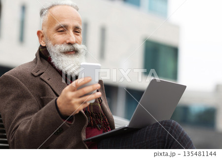 Senior grey-haired businessman with laptop sitting on bench next to his office and using smartphone, chatting with clients, copy space. Elderly man using modern laptop and checking messages on phone Senior grey-haired businessman with laptop sitting on bench next to his office and using smartphone, chatting with clients, copy space. Elderly man using modern laptop and checking messages on phone 135481144