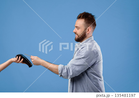 Side view of smiling young man paying with credit card on payment terminal, blue studio background. Millennial guy shopping contactless, using electronic money during covid outbreak 135481197
