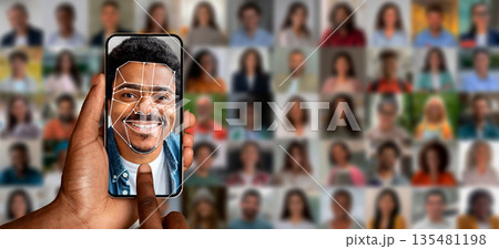 A hand holds a smartphone displaying a face recognition scan of a smiling man. Behind are various blurred portraits of people, illustrating advanced identification technology in action. 135481198