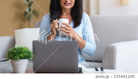 Technology And People. Attractive Mature Lady With Long Dark Hair Working Remotely On Laptop At Home Office, Enjoying Morning Coffee, Woman Sitting On The Couch With Computer On Desk Technology And People. Attractive Mature Lady With Long Dark Hair Working Remotely On Laptop At Home Office, Enjoying Morning Coffee, Woman Sitting On The Couch With Computer On Desk 135481200