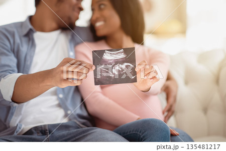 Positive black couple showing ultrasound photo of their unborn baby at home, selective focus on hands. Happy expectant family with sonogram of their child, creating happy memories together 135481217