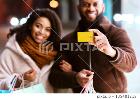 Consumerism And Relationship. Portrait of smiling black man showing debit credit card to camera, woman embracing him and holding shopping bags, family near mall in the evening, blurred background Consumerism And Relationship. Portrait of smiling black man showing debit credit card to camera, woman embracing him and holding shopping bags, family near mall in the evening, blurred background 135481218