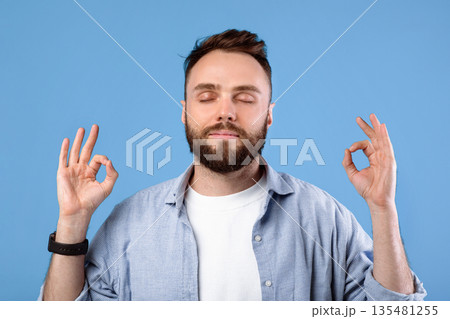 Calm young man meditating to relieve stress, finding inner peace and harmony on blue studio background. Bearded millennial guy practicing mindfulness and deep concentration 135481255
