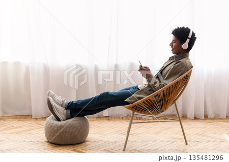 Black teenager relaxing in modern armchair, enjoying music on smartphone at home, banner design with copy space. African American youth in headphones listening to his favorite song Black teenager relaxing in modern armchair, enjoying music on smartphone at home, banner design with copy space. African American youth in headphones listening to his favorite song 135481296
