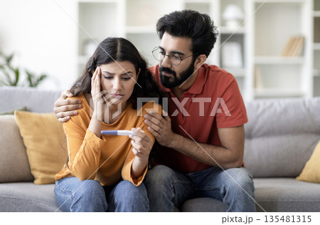 Infertility Problems. Portrait Of Upset Young Indian Couple Looking At Negative Pregnancy Test While Sitting Together On Couch At Home, Caring Eastern Husband Comforting Depressed Wife, Free Space Infertility Problems. Portrait Of Upset Young Indian Couple Looking At Negative Pregnancy Test While Sitting Together On Couch At Home, Caring Eastern Husband Comforting Depressed Wife, Free Space 135481315