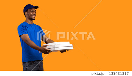 Fast Delivery Service. Cheerful african american courier wearing blue cap and uniform holding pizza boxes from restaurant and giving it to free copy space, yellow studio background, banner, panorama 135481317