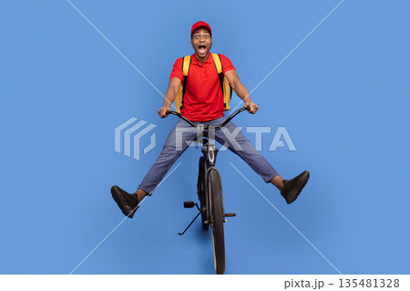 Fast Delivery Service. Funny african american courier guy in red cap and uniform riding bicycle and wearing thermo backpack bag isolated on blue studio background. Shipment service during lockdown Fast Delivery Service. Funny african american courier guy in red cap and uniform riding bicycle and wearing thermo backpack bag isolated on blue studio background. Shipment service during lockdown 135481328