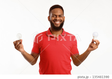 African American Guy Holding Two Light Bulbs Standing On White Studio Background, Choosing Energy-Saving Lightbulb. Save Energy, Safe Light-Bulbs Disposal Concept African American Guy Holding Two Light Bulbs Standing On White Studio Background, Choosing Energy-Saving Lightbulb. Save Energy, Safe Light-Bulbs Disposal Concept 135481390
