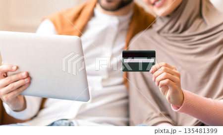 Muslim Couple Shopping Online With Credit Card And Digital Tablet Computer Sitting On Couch At Home, Focus On Bank Card. Happy Customers, Internet Shop Sales And Discounts Concept. Shallow Depth 135481399