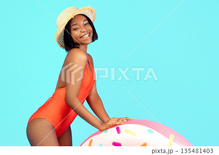 Full length portrait of joyful black lady in swimming suit with donut shaped inflatable ring on blue studio background. Gorgeous fit young lady in bikini posing with pool float, having summer vacation 135481403