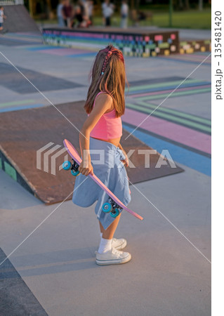 A preschool-age girl with a skateboard in her hands at sunset on a late summer evening. 135481420