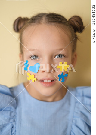 Portrait of a girl with clear eyes, ornaments on her cheek in the shape of hearts and pieces of yellow and blue puzzles. Awareness and social support for children with down syndrome Portrait of a girl with clear eyes, ornaments on her cheek in the shape of hearts and pieces of yellow and blue puzzles. Awareness and social support for children with down syndrome 135481532