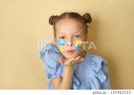 Portrait of a girl with ornaments on her cheek in the shape of hearts and puzzles in yellow and blue. The child sends an air kiss to the world full of kindness and love. World Down Syndrome Day 135481533