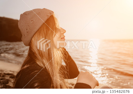 Woman, sunset, beach. Young blonde girl wearing a beanie hat sitting by the ocean, enjoying the serene golden hour light. 135482076