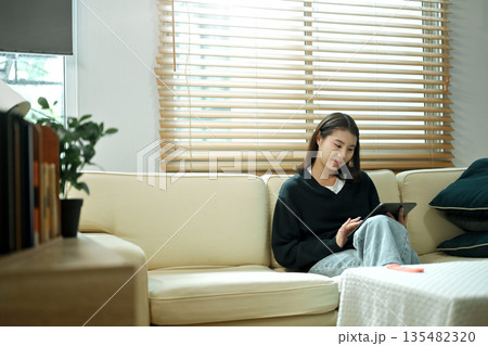 A young woman relaxing on a sofa using a tablet at home, enjoying a calm holiday mood with comfort and leisure time A young woman relaxing on a sofa using a tablet at home, enjoying a calm holiday mood with comfort and leisure time 135482320