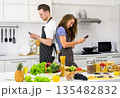 Young caucasian couple in aprons standing back to back in kitchen at home. Man and woman using mobile phone in front of fresh fruits and vegetables arrange on white countertop 135482832