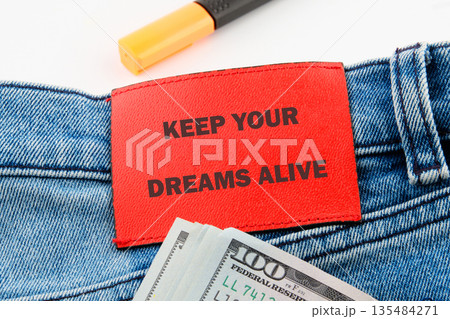 red rectangular label with the words keep your dreams alive rests on blue jeans with stacks of one hundred United States dollar bills, and a highlighter red rectangular label with the words keep your dreams alive rests on blue jeans with stacks of one hundred United States dollar bills, and a highlighter 135484271