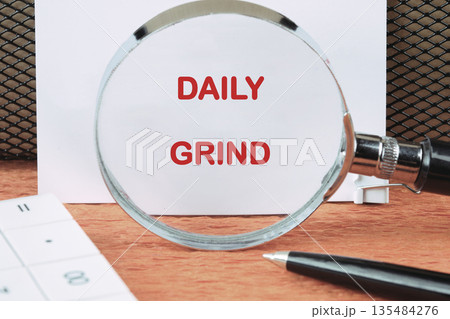 A magnifying glass enlarges the words Daily Grind on a white sheet of paper. A pen and calculator rest nearby on a wooden surface in an office setting 135484276