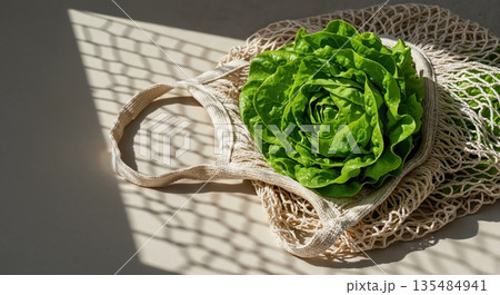 A vibrant green head of fresh butter lettuce rests inside an eco-friendly beige string bag on a light surface, illuminated by natural sunlight creating a patterned shadow. 135484941