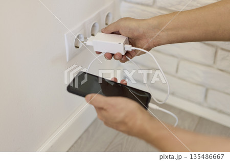 Hands plugging phone charger into wall socket with adapter and cable visible Hands plugging phone charger into wall socket with adapter and cable visible 135486667