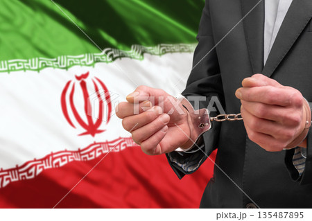 A man in a business suit stands in handcuffs against the flag of Iran. A man in a business suit stands in handcuffs against the flag of Iran. 135487895